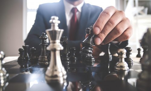 Retro style image of a businessman with clasped hands planning strategy with chess figures on an old wooden table.
